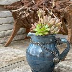 Une composition végétale de plantes succulentes avec un pichet artisan en céramique bleu et un sautoir en métal maillon chainette et frange