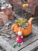 Composition végétale de plantes succulentes avec une théière en céramique orange et un personnage porte photos en résine rose et vert