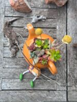 Composition végétale de plantes succulentes avec une théière en céramique orange et un personnage porte photos en résine rose et vert